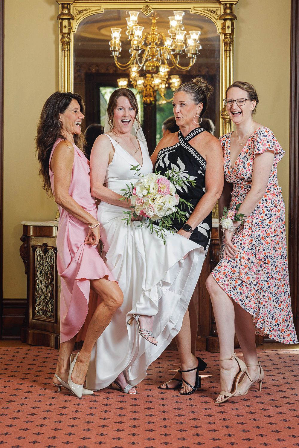 Wedding party laughs together in elegant venue with floral dresses, chandelier and patterned carpet.