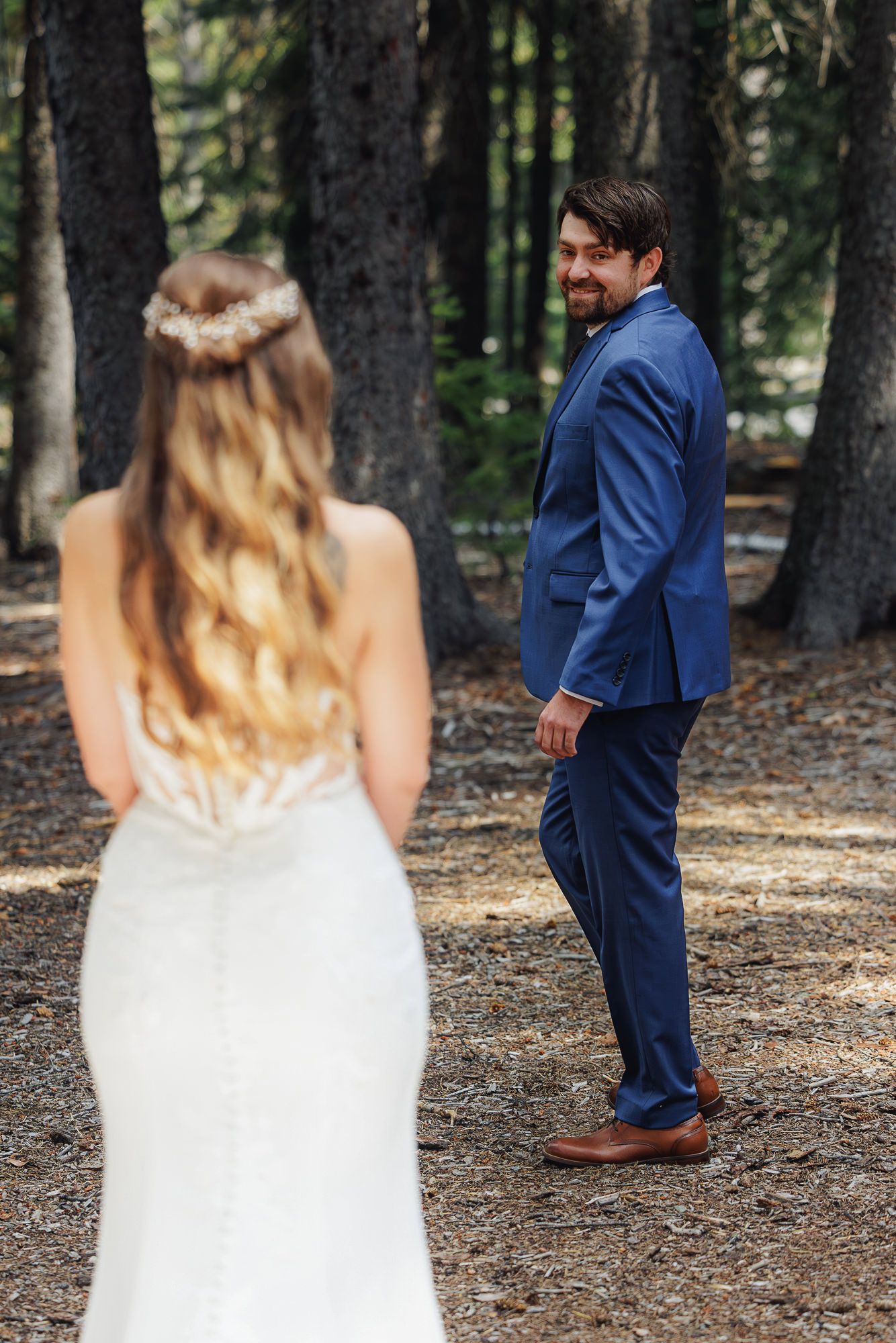 A bride in a white dress and groom in a navy suit share their first look moment in a wooded forest setting.