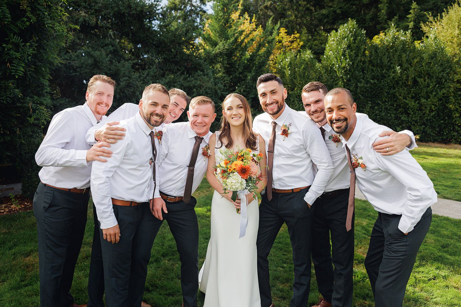 Wedding party members share joyful embraces and laughter while posing outdoors in formal attire on a sunny day.