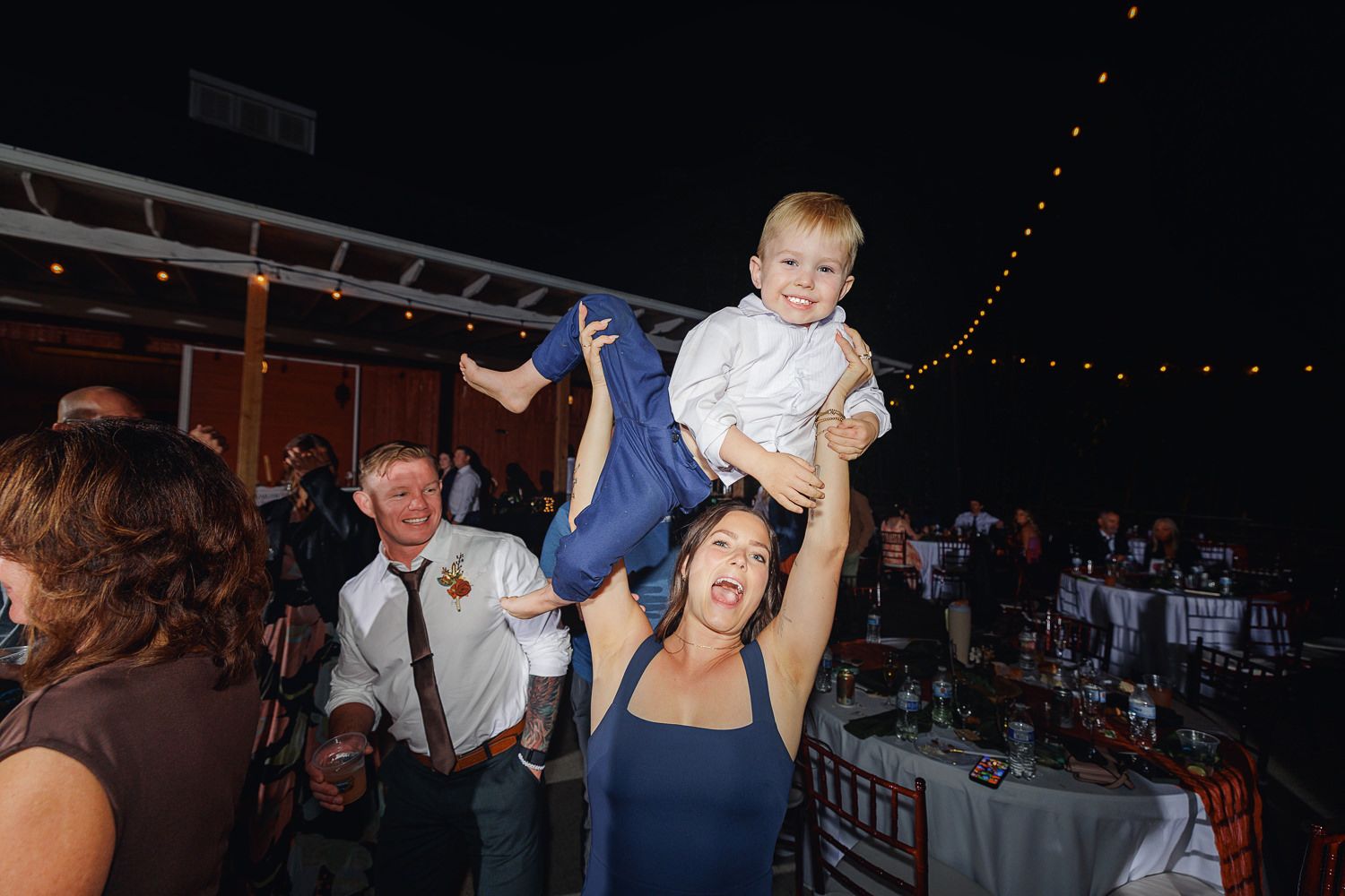 Wedding guests dance and celebrate on a dimly lit dance floor with string lights overhead during an evening reception.