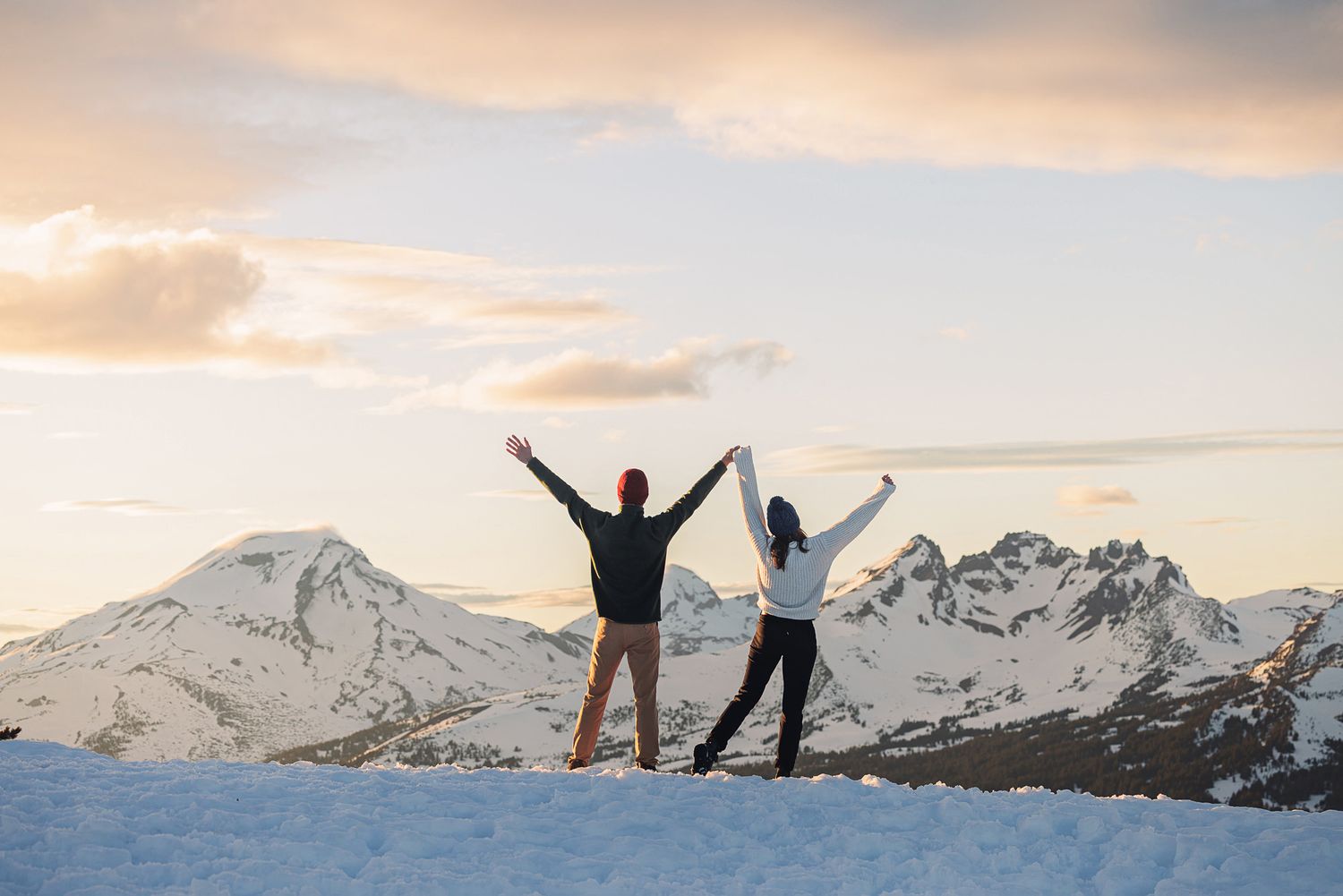 Couple raising hands together on snowy mountain summit at sunset with snow-capped peaks in background.