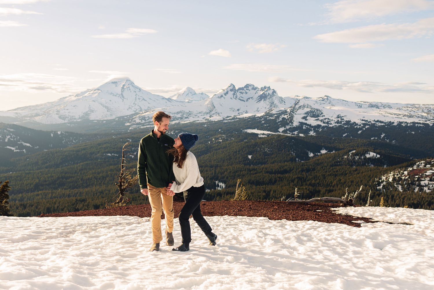 Couple walking hand in hand on snowy mountain summit with stunning Cascade Range peaks and evergreen forest backdrop.