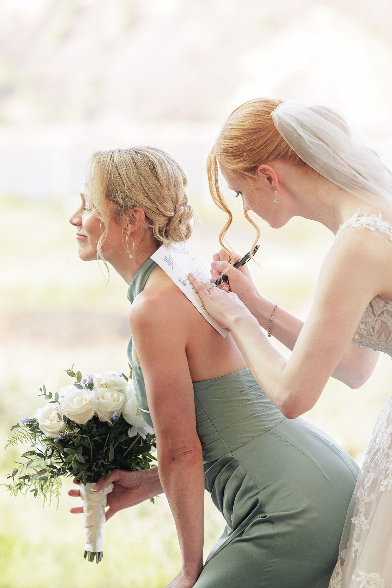 Bride signing the back of a bridesmaid in a sage green dress holding white roses at an outdoor wedding.