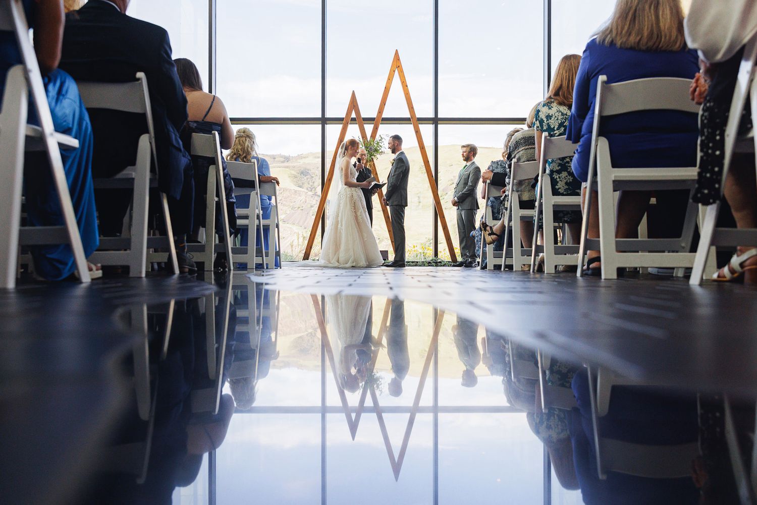 Elegant wedding ceremony with couple at altar, wood triangle arch, floor-to-ceiling windows, and guests seated in white chairs.