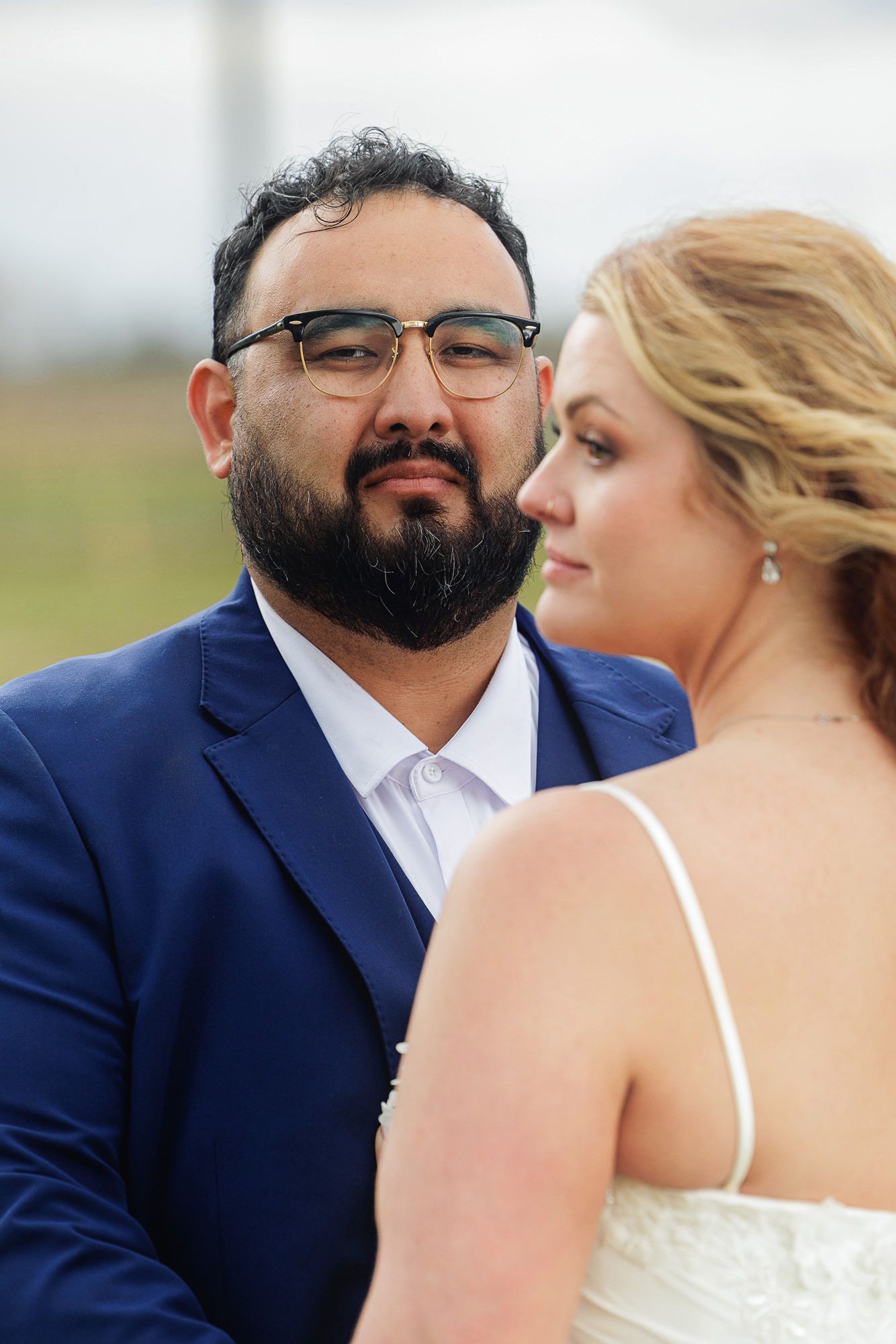 A bearded groom in a blue suit and glasses gazes lovingly at his bride in a white wedding dress outdoors.