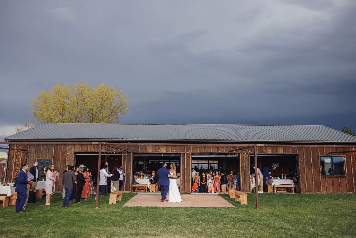 Outdoor wedding ceremony at a rustic wooden barn venue under dramatic stormy skies with guests gathered on green lawn.