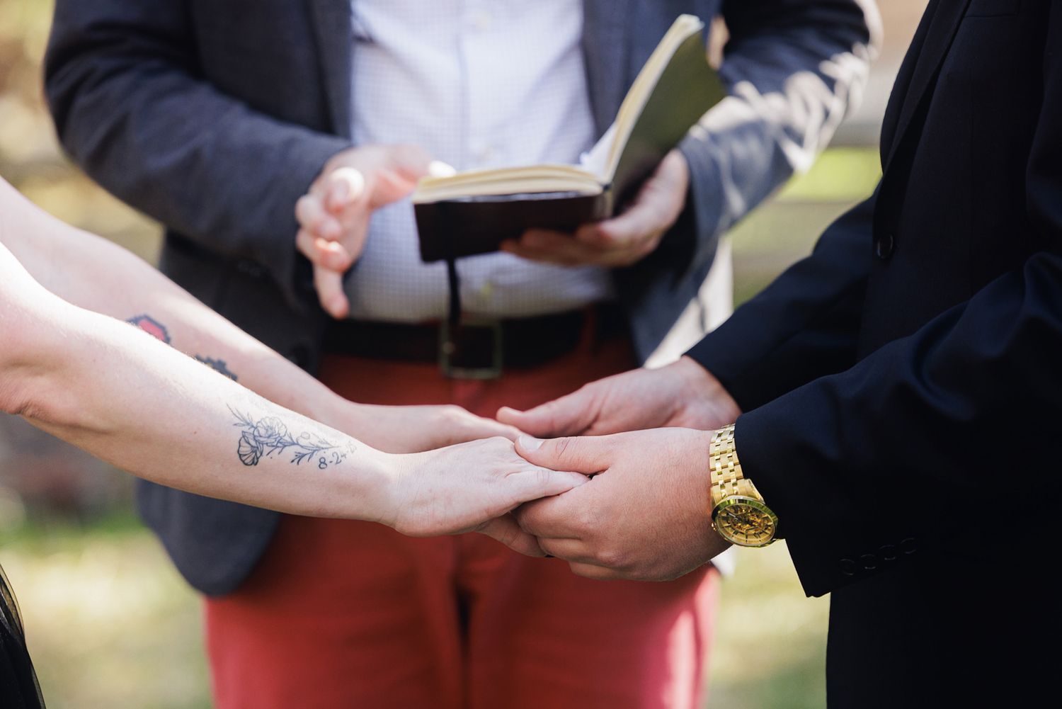 Couple holding hands during wedding ceremony with officiant holding book in background, bride has floral tattoo on arm.