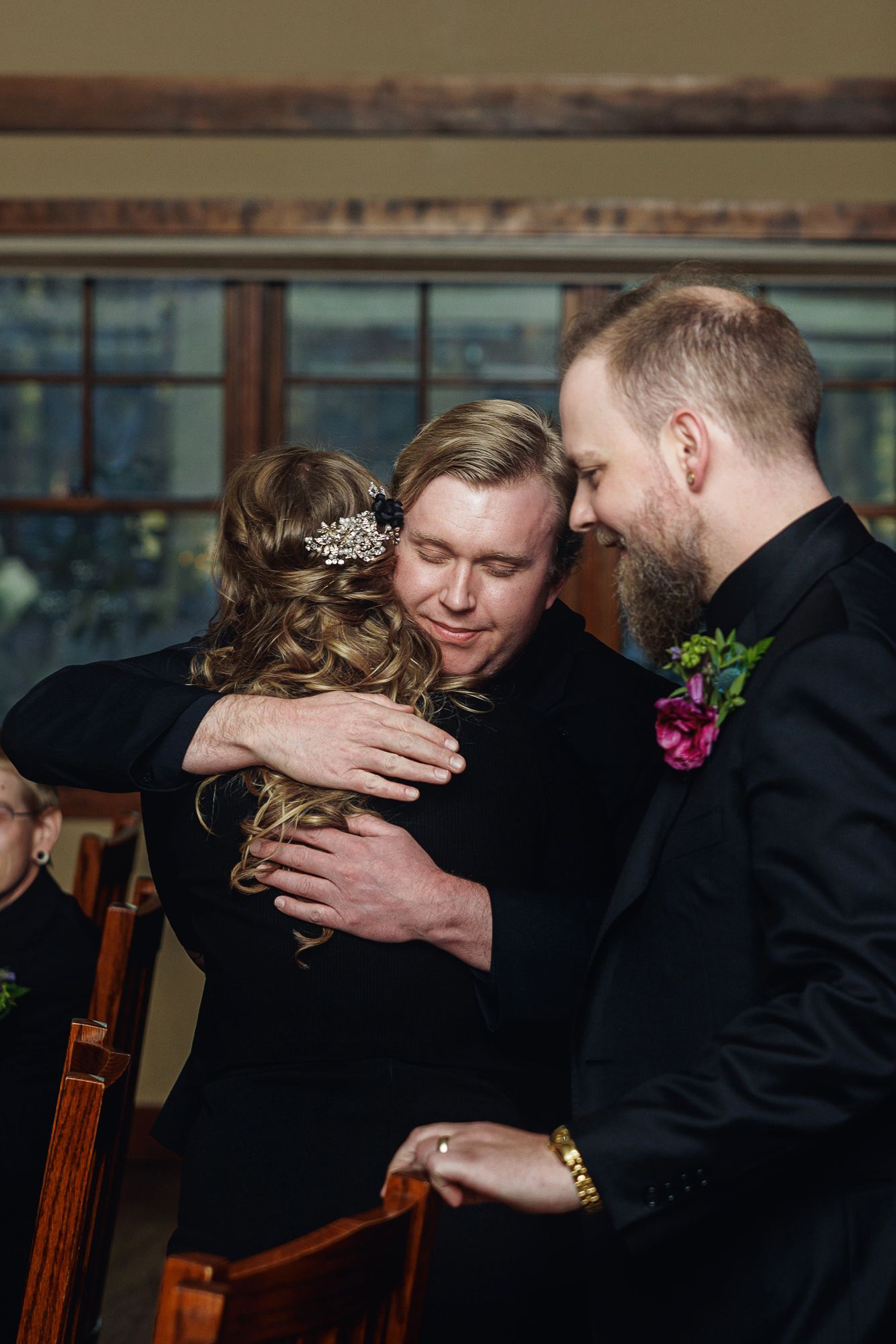 Two grooms in black suits share an emotional hug with a woman in a hair accessory at a wedding ceremony.