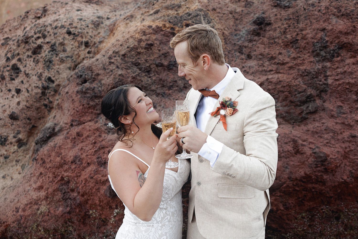 Bride in a white dress and groom in a cream suit with a floral boutonniere share a joyful toast with champagne against red volcanic cliffs.