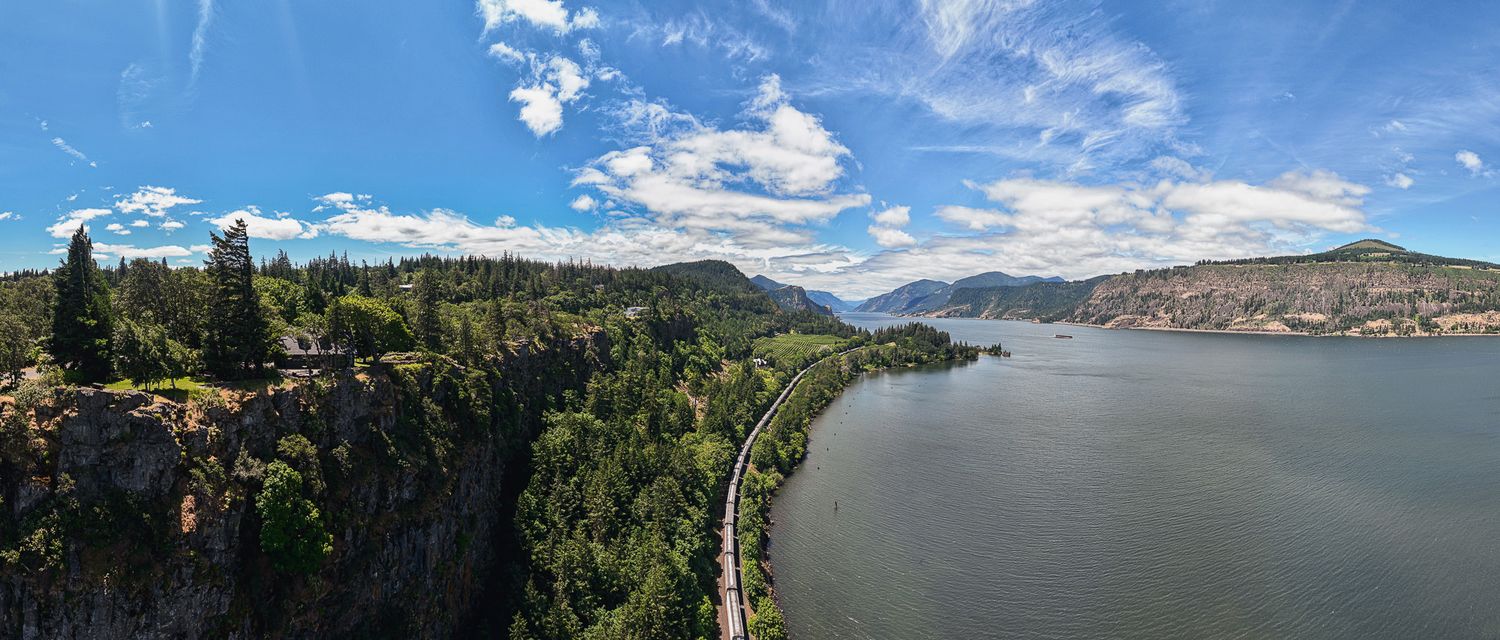 Panoramic view of Columbia River Gorge with lush green cliffs, railway, and blue sky with clouds.