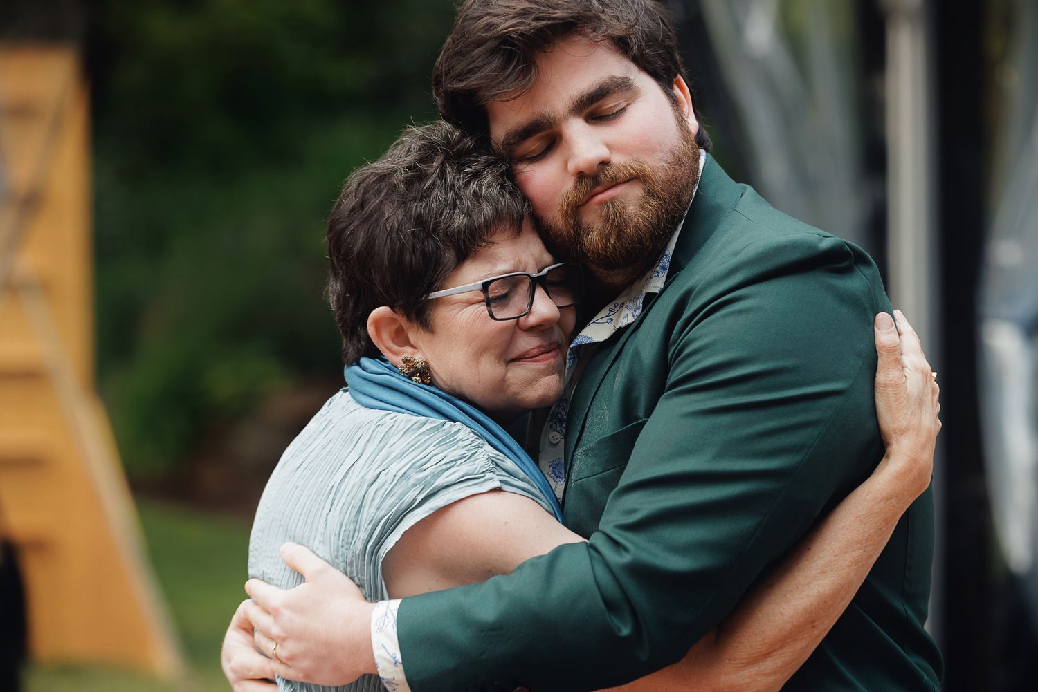 A young bearded man in a green suit shares a warm embrace with an older woman wearing glasses outdoors.