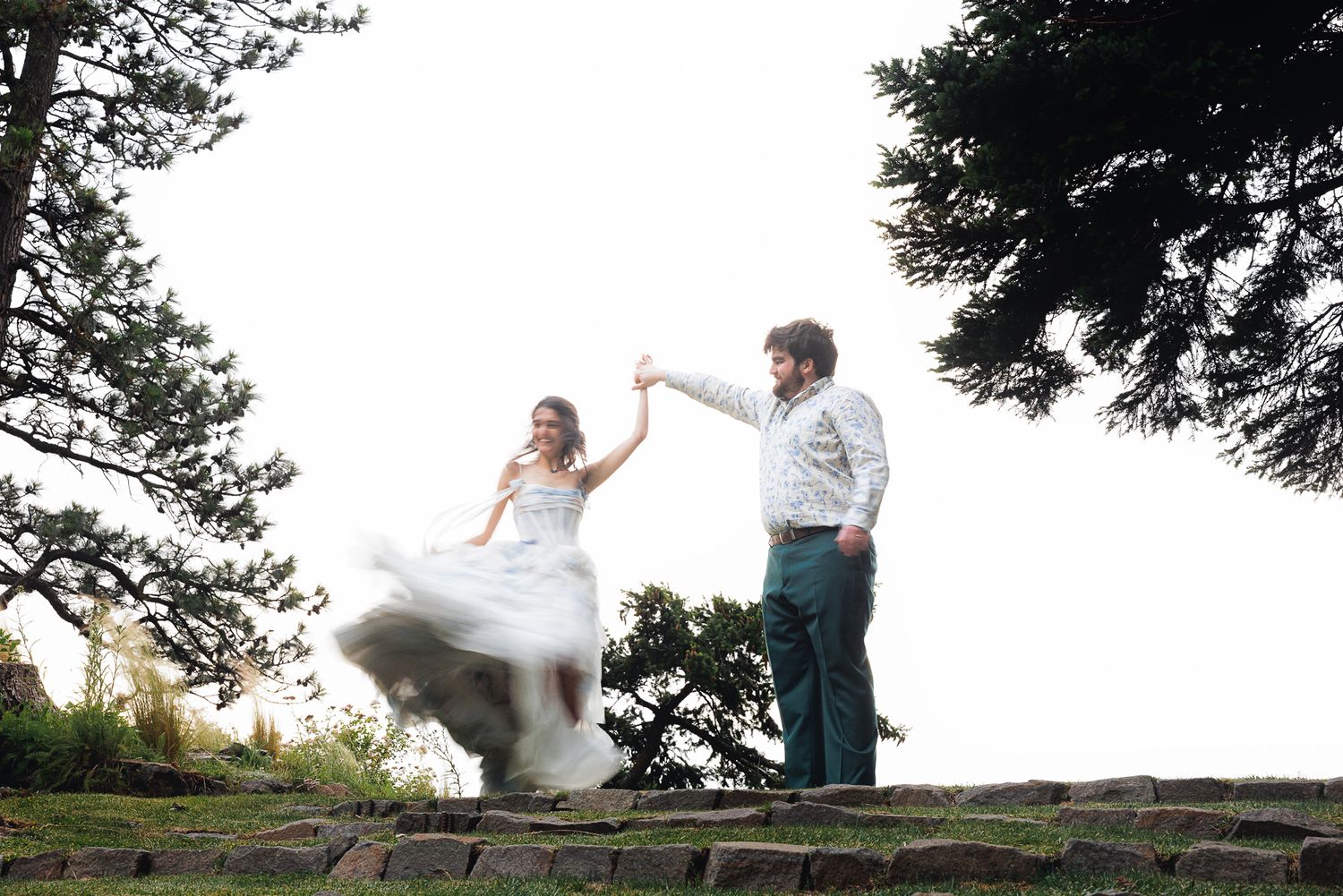 Bride and groom dancing outdoors on stone steps surrounded by tall trees on their wedding day.