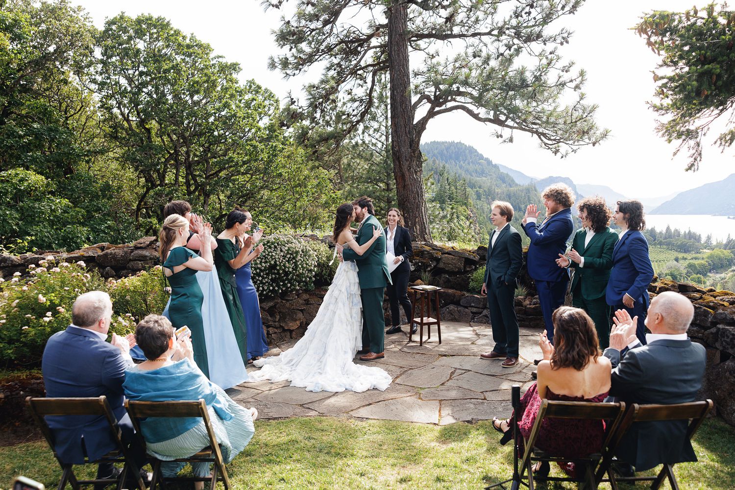 Outdoor wedding ceremony on a stone patio with mountain views, bride and groom kissing surrounded by guests in teal and blue attire.