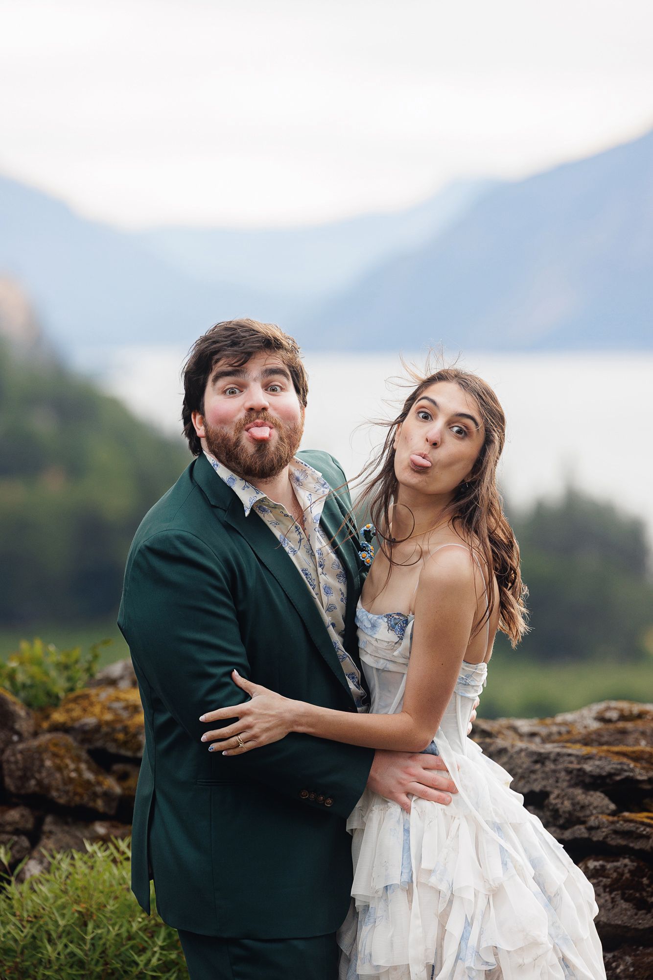 A playful bride and groom making funny faces outdoors with a scenic mountain lake backdrop on their wedding day.