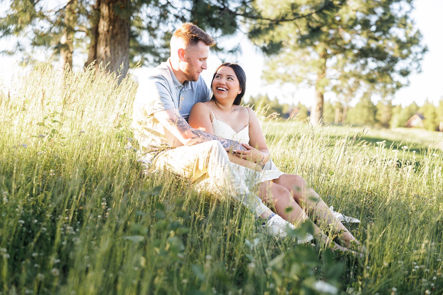 Couple laughing together in tall green grass near pine trees during golden hour outdoor engagement session.