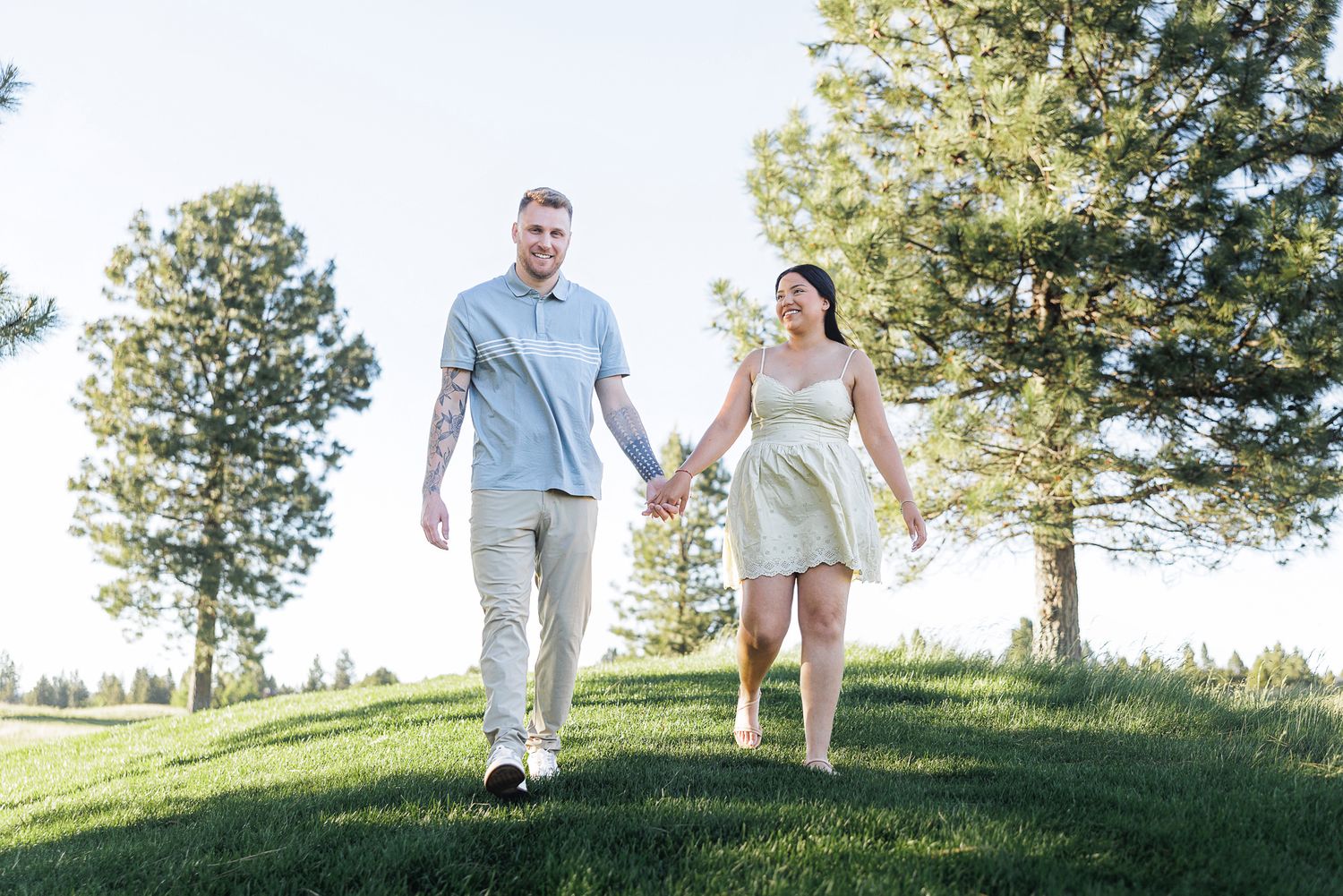 Couple holding hands walking on green grass hill with tall pine trees in background on sunny day.