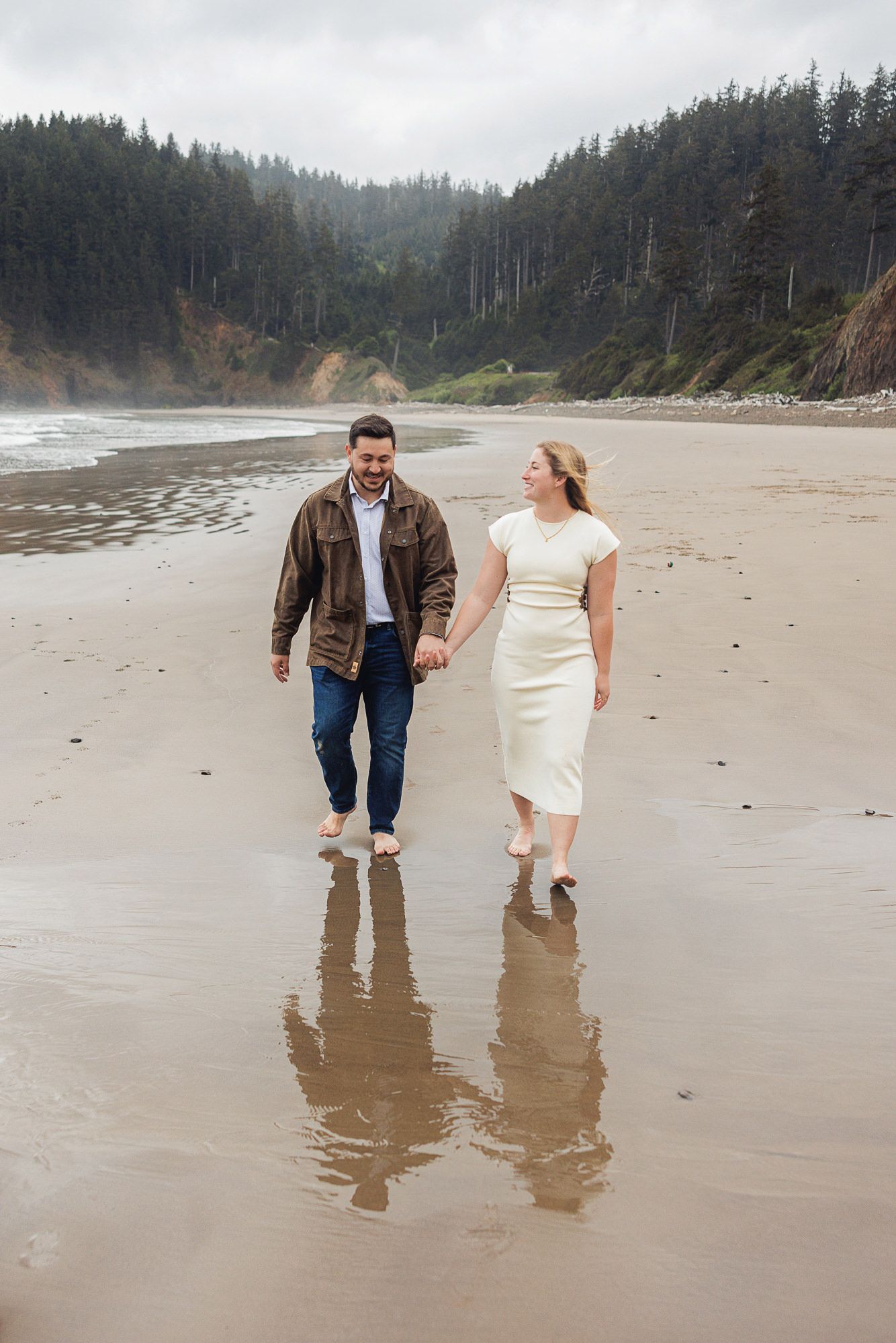 Couple walks hand-in-hand barefoot on a misty Pacific Northwest beach, woman in white dress, man in brown leather jacket.
