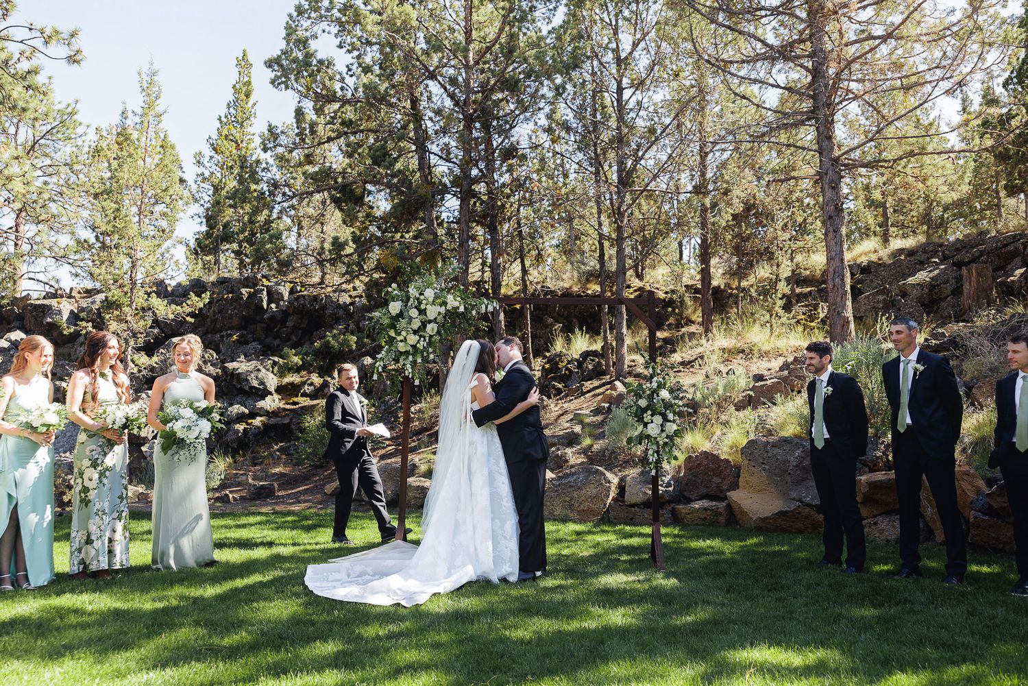 Outdoor wedding ceremony on green lawn with bride and groom at floral arch, wedding party, and pine trees in background.