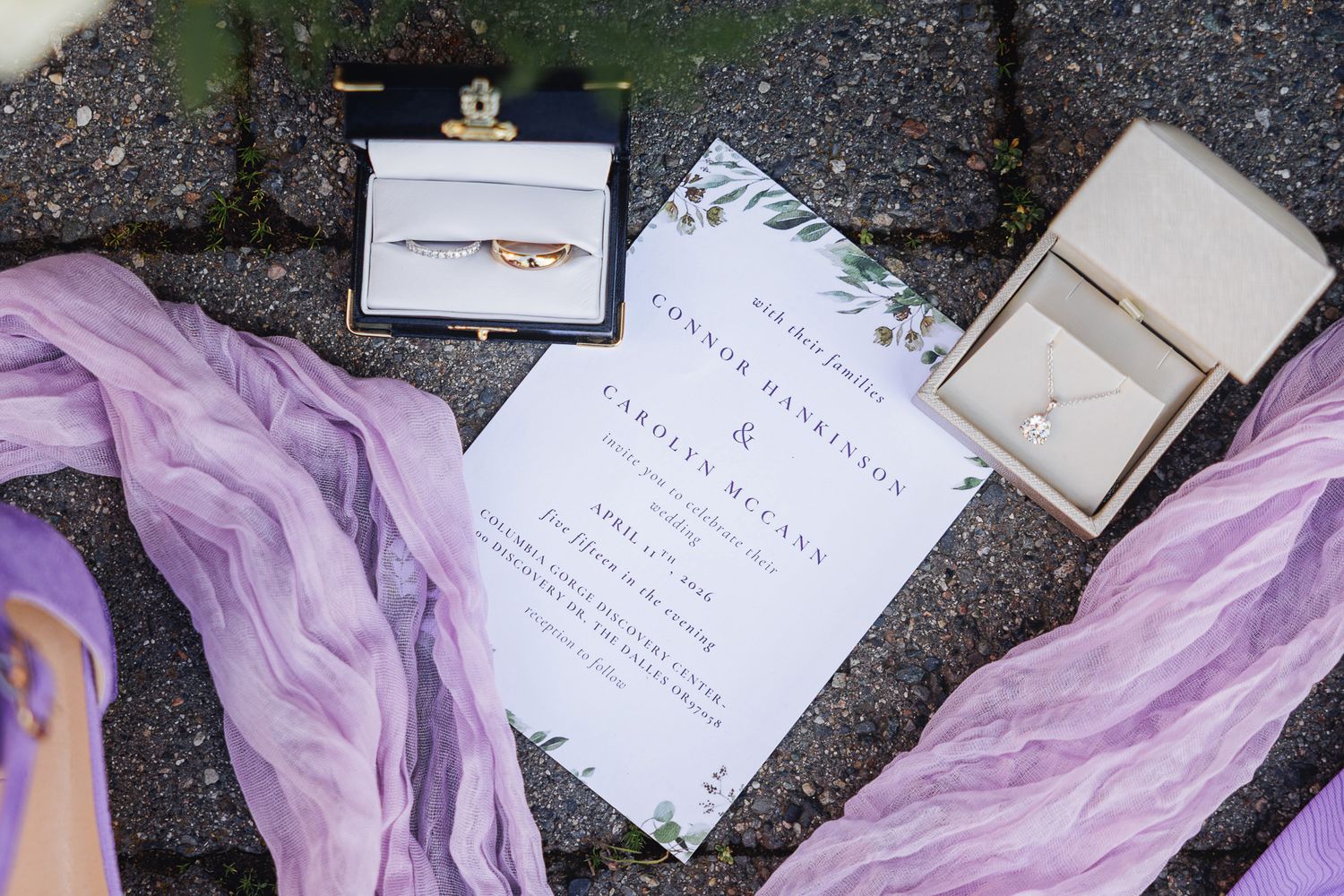 Elegant wedding invitation with greenery accents, gold rings in velvet boxes, and purple silk ribbon on stone pavement.