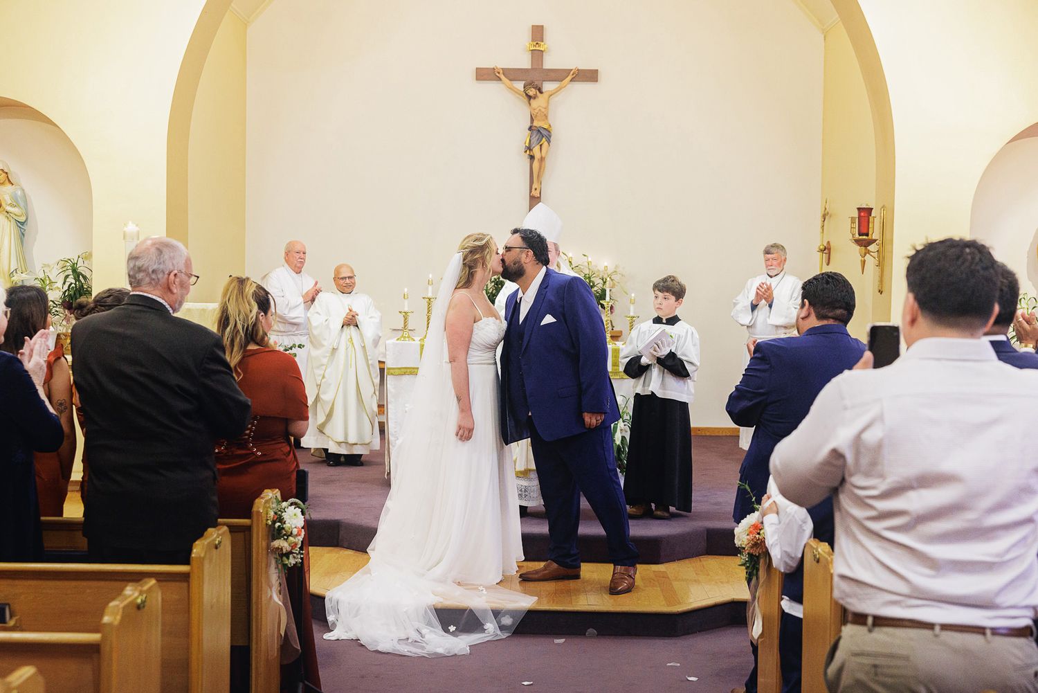 Bride and groom share a kiss at the altar during a Catholic church wedding ceremony with guests watching.