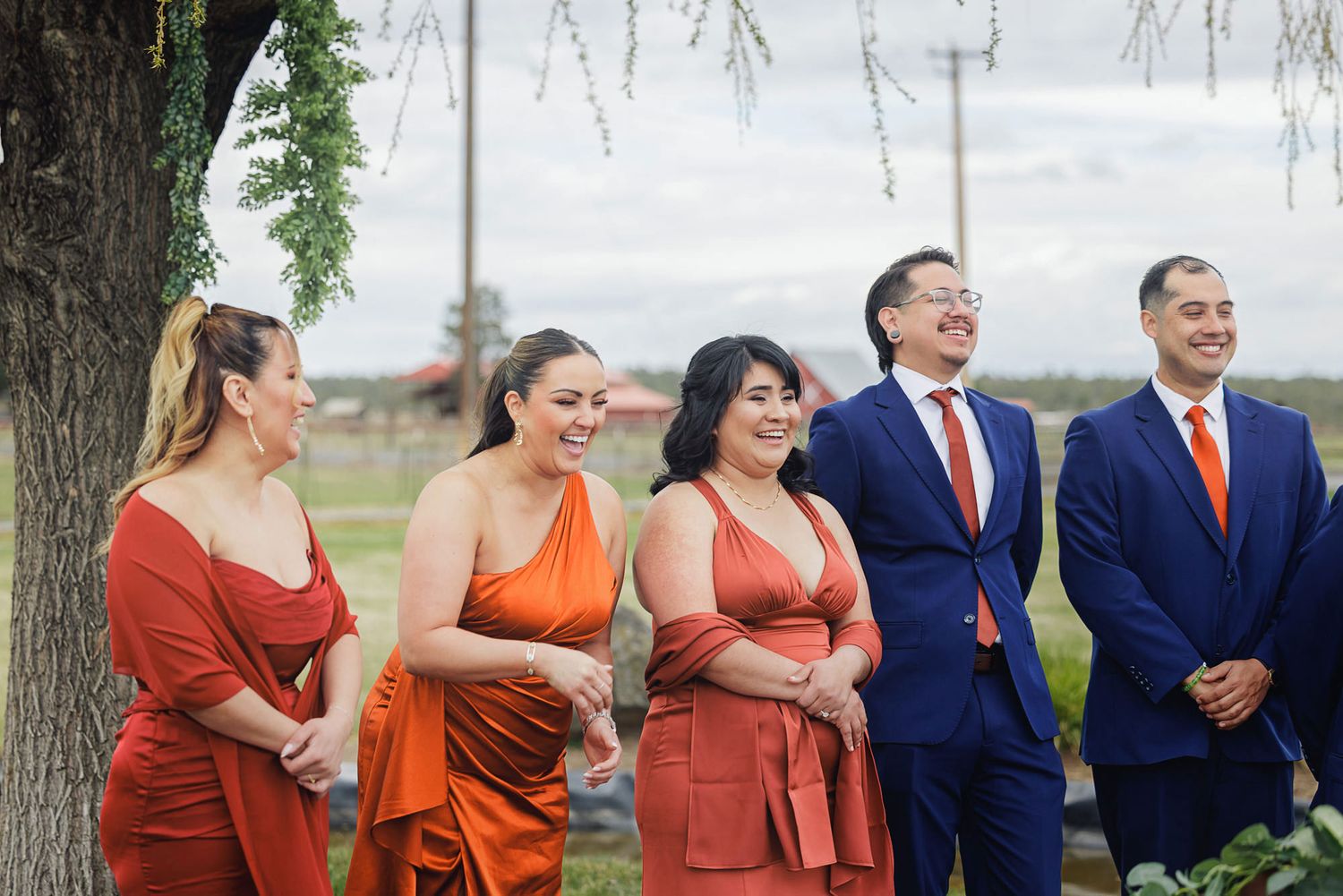 Wedding party in orange dresses and navy suits with orange ties smiling outdoors near a tree.