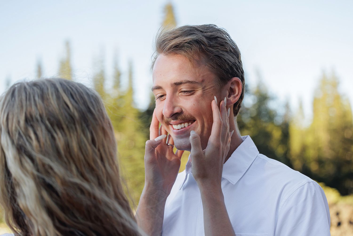 Woman tenderly touching smiling man's face outdoors with evergreen trees in background on sunny day.