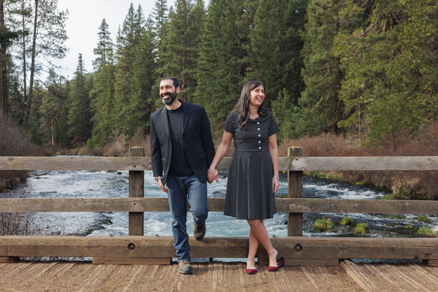 Couple holding hands on a wooden bridge surrounded by pine trees and a flowing river in a forest setting.