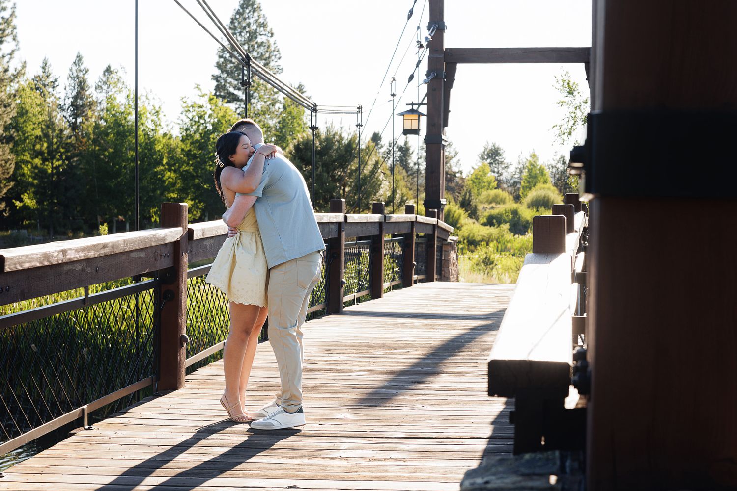 Couple sharing a romantic kiss on a sunny wooden boardwalk surrounded by lush green trees.