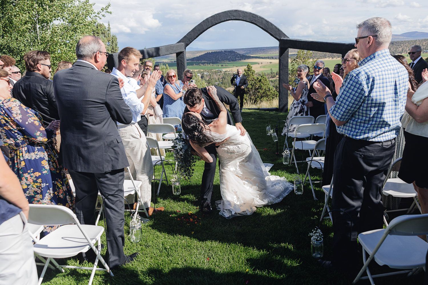 An outdoor wedding ceremony takes place under an arch with scenic mountain views in the background on a sunny day.