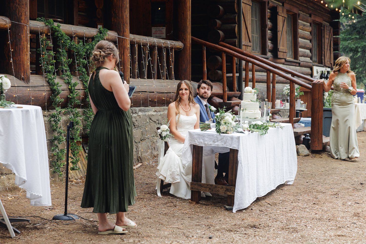 A group gathers at a rustic outdoor venue with white-clothed tables and wooden structures in a forest setting.