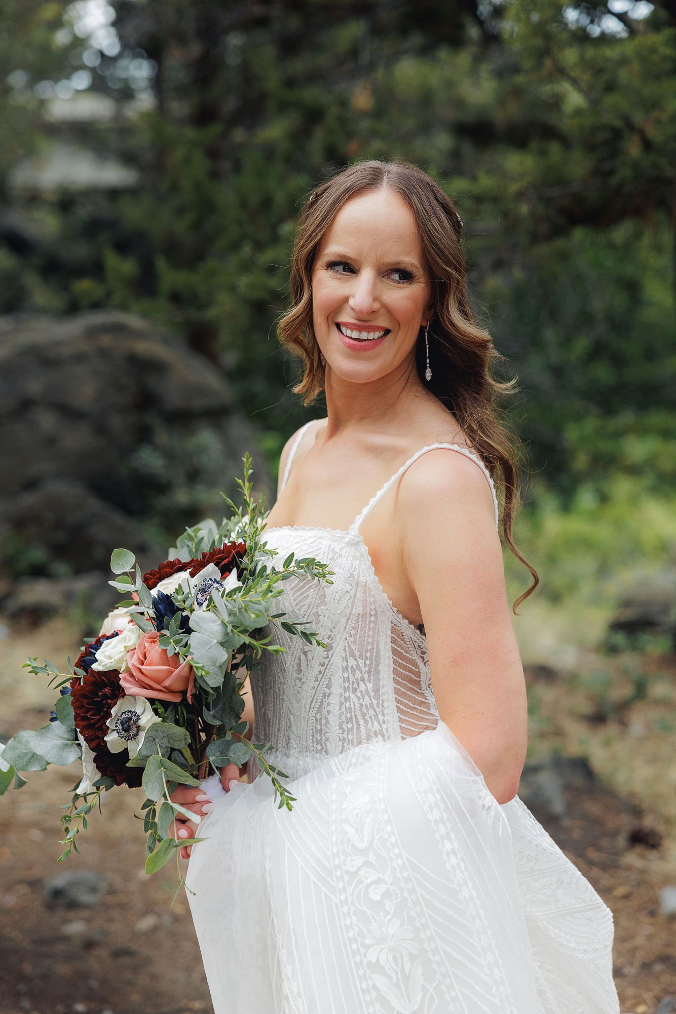 A bride in a white wedding dress holds a romantic bouquet of pink roses and greenery against a natural outdoor backdrop.