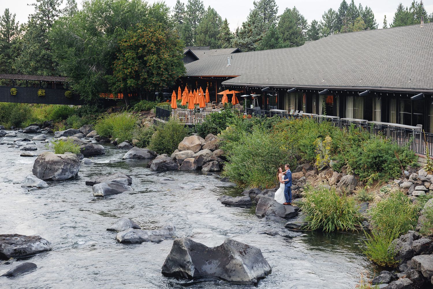 A scenic river with large boulders and rocks flows past rustic buildings nestled among green trees along the riverbank.