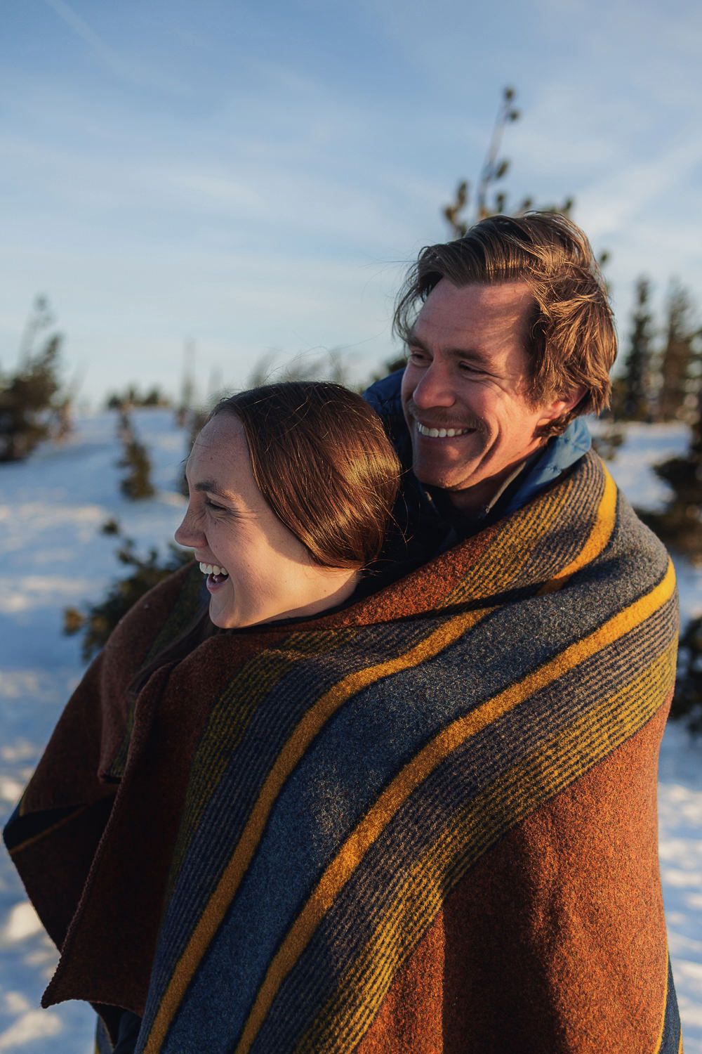 A couple laughs and snuggles together in winter snow with evergreen trees in the background.