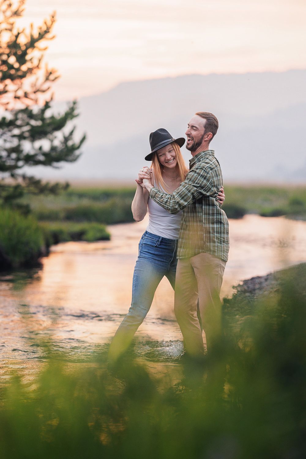 A couple embraces while dancing together at sunset by a tranquil lake surrounded by mountains and greenery.