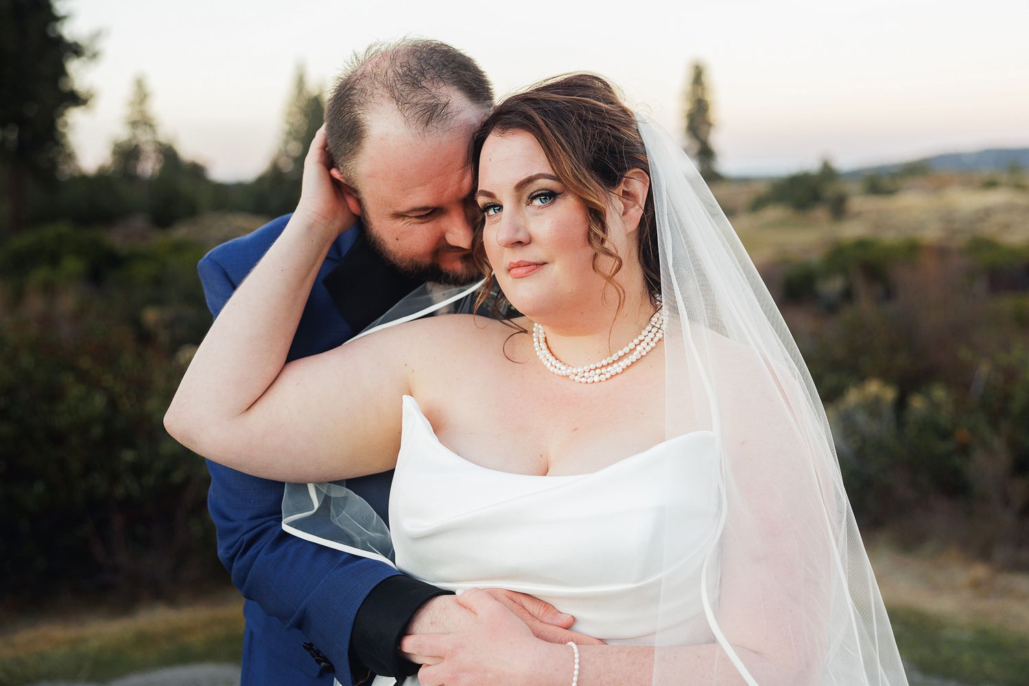 A couple in wedding attire shares an intimate moment together in a scenic outdoor setting at sunset.