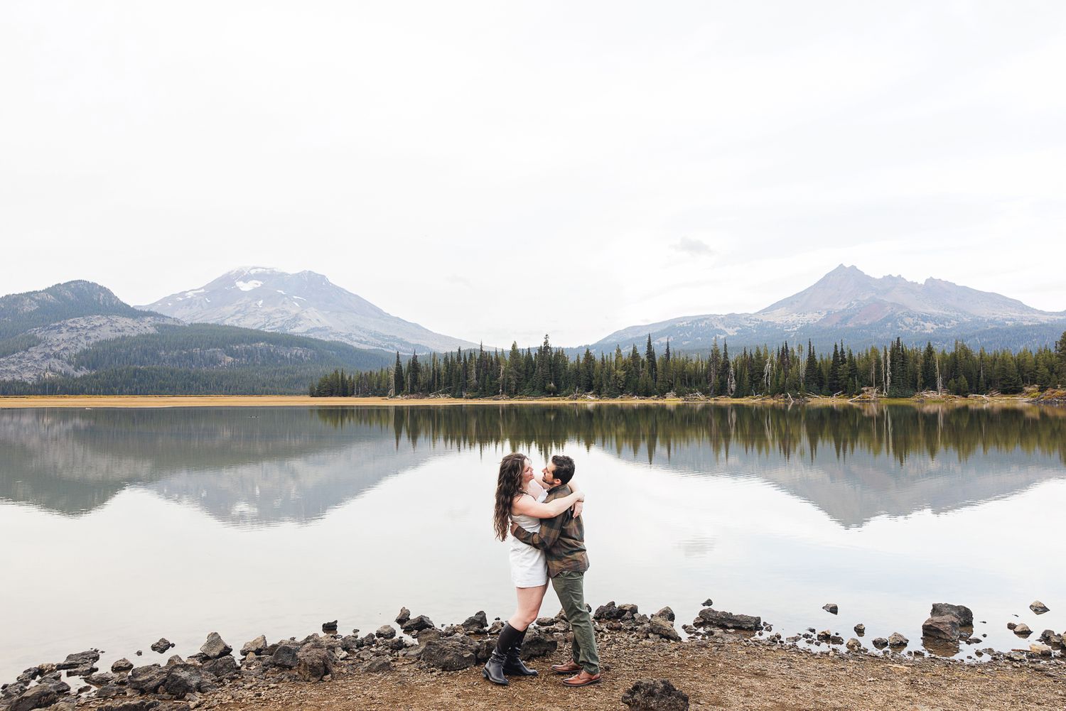 A couple embraces by a serene mountain lake with snow-capped peaks reflected in the still water.