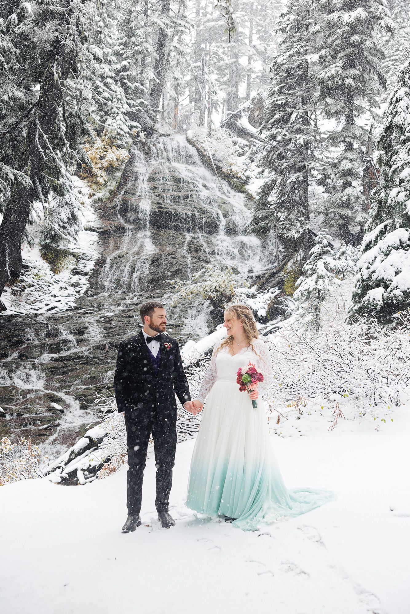 A couple shares a romantic moment in a snow-covered forest with evergreen trees and white winter scenery.