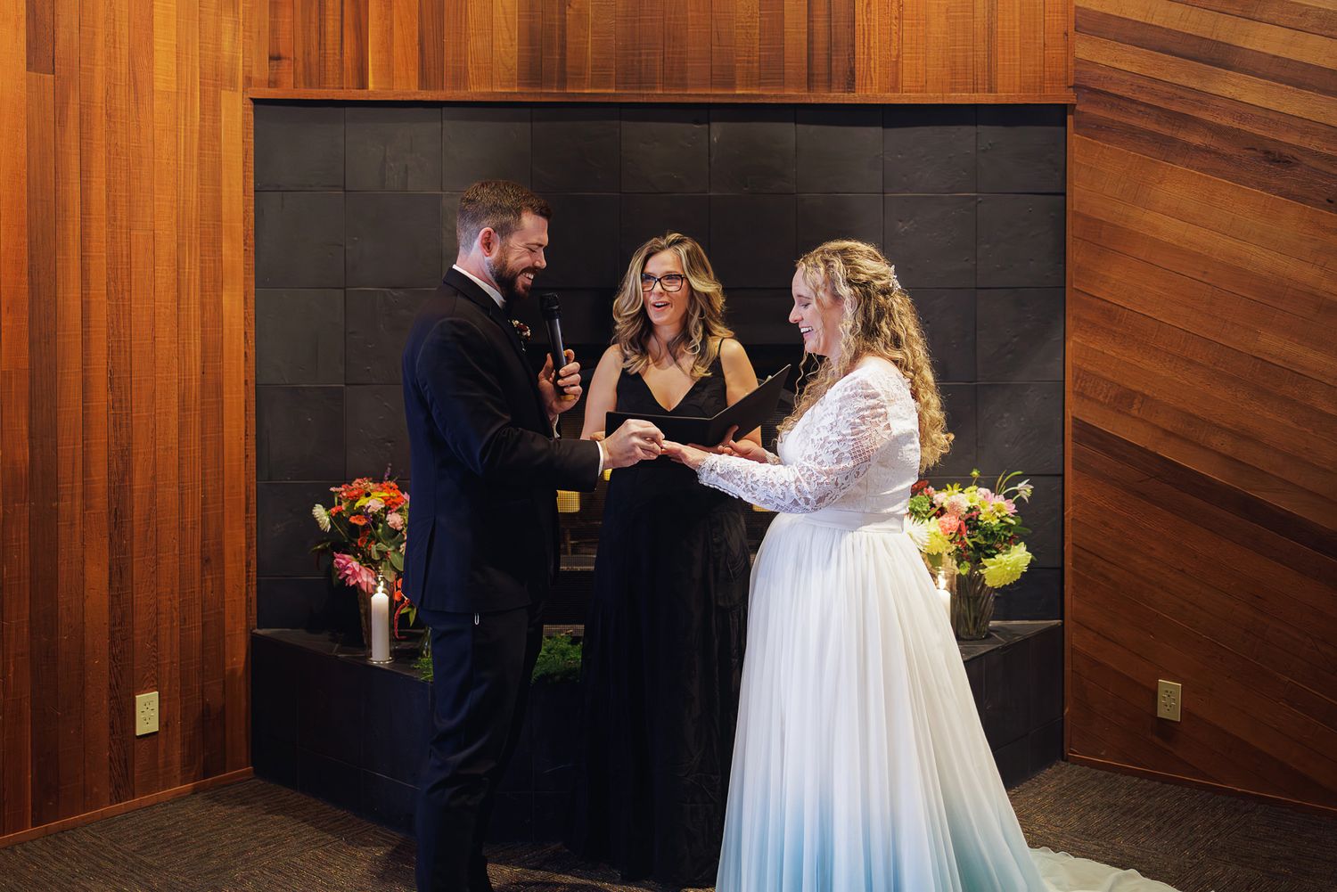 A wedding ceremony takes place in a wood-paneled room with floral arrangements on either side.