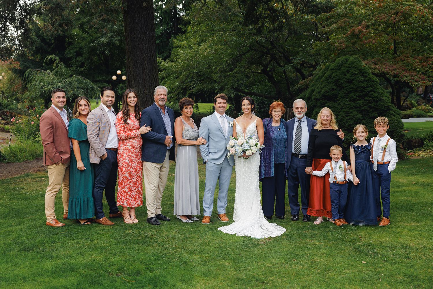 A wedding party and guests gather outdoors in a garden setting wearing formal attire and colorful dresses.