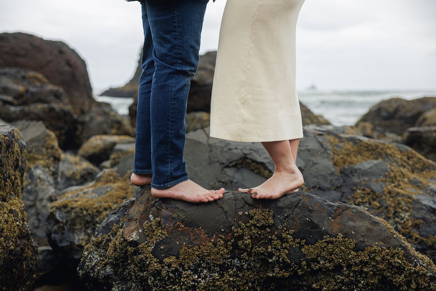 Barefoot couple standing on mossy coastal rocks near the ocean, one in jeans and one in a cream skirt.