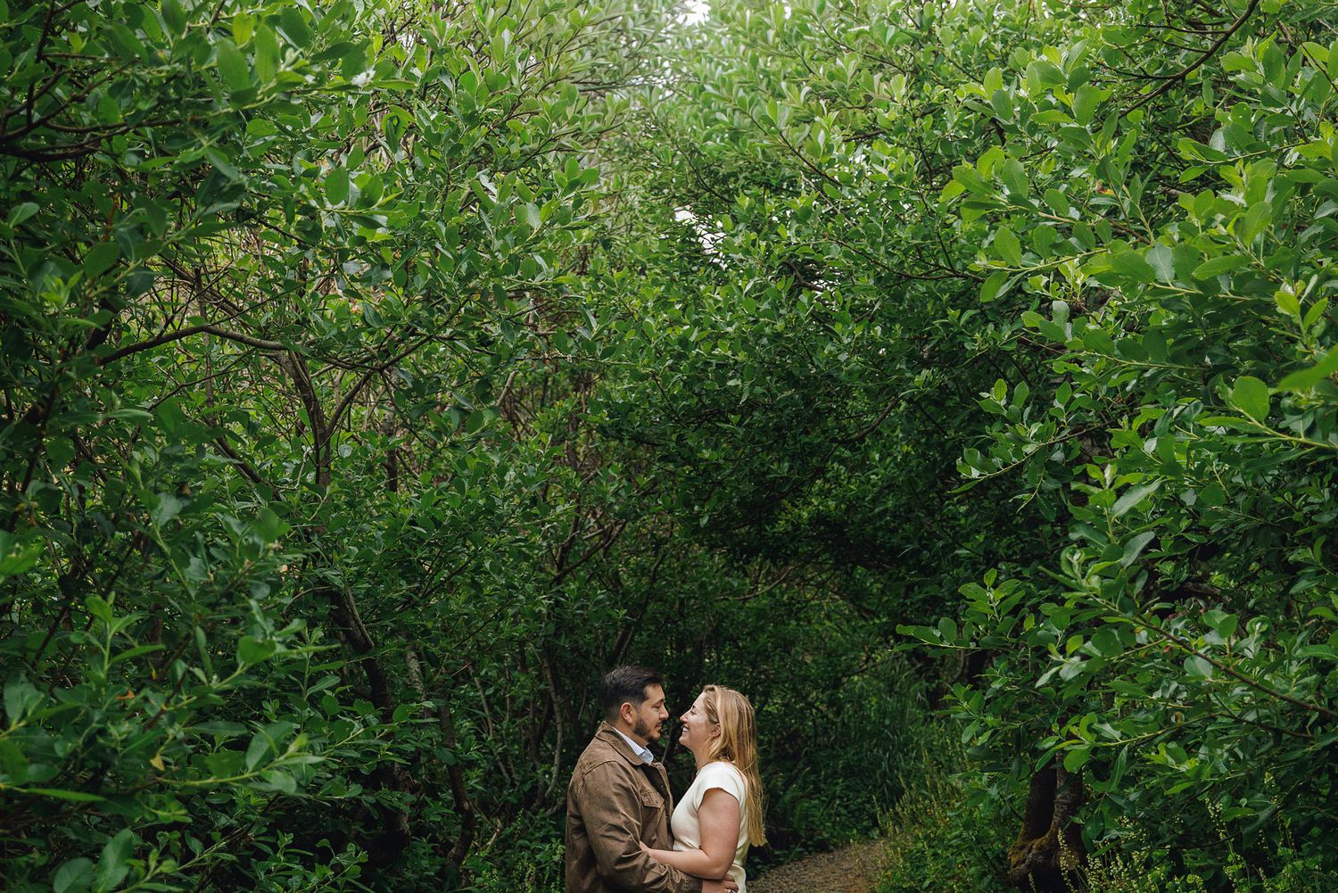 Couple sharing a romantic moment surrounded by lush green foliage in a garden pathway.