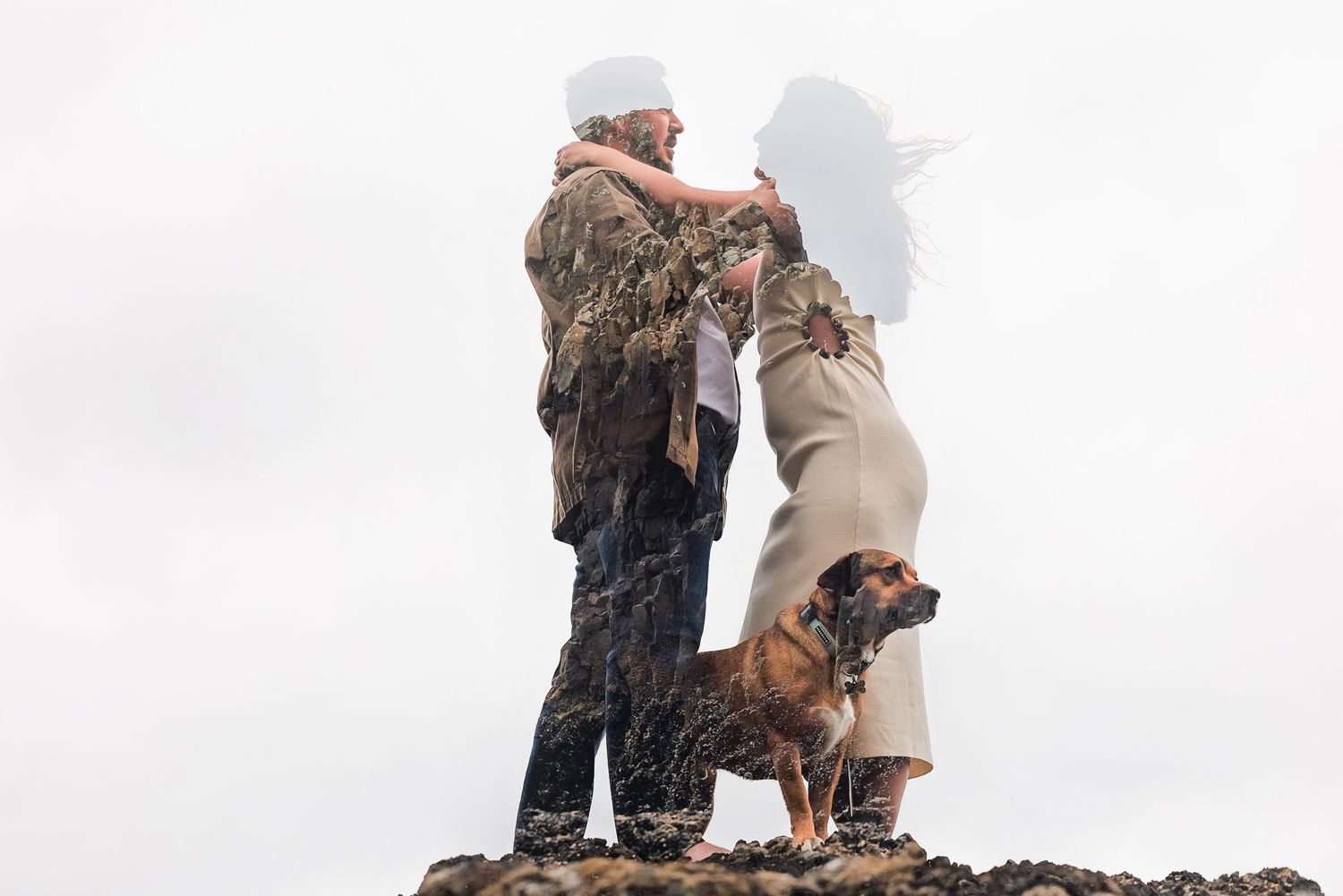 Couple embracing on rocky terrain with a brown dog beside them against a cloudy sky.
