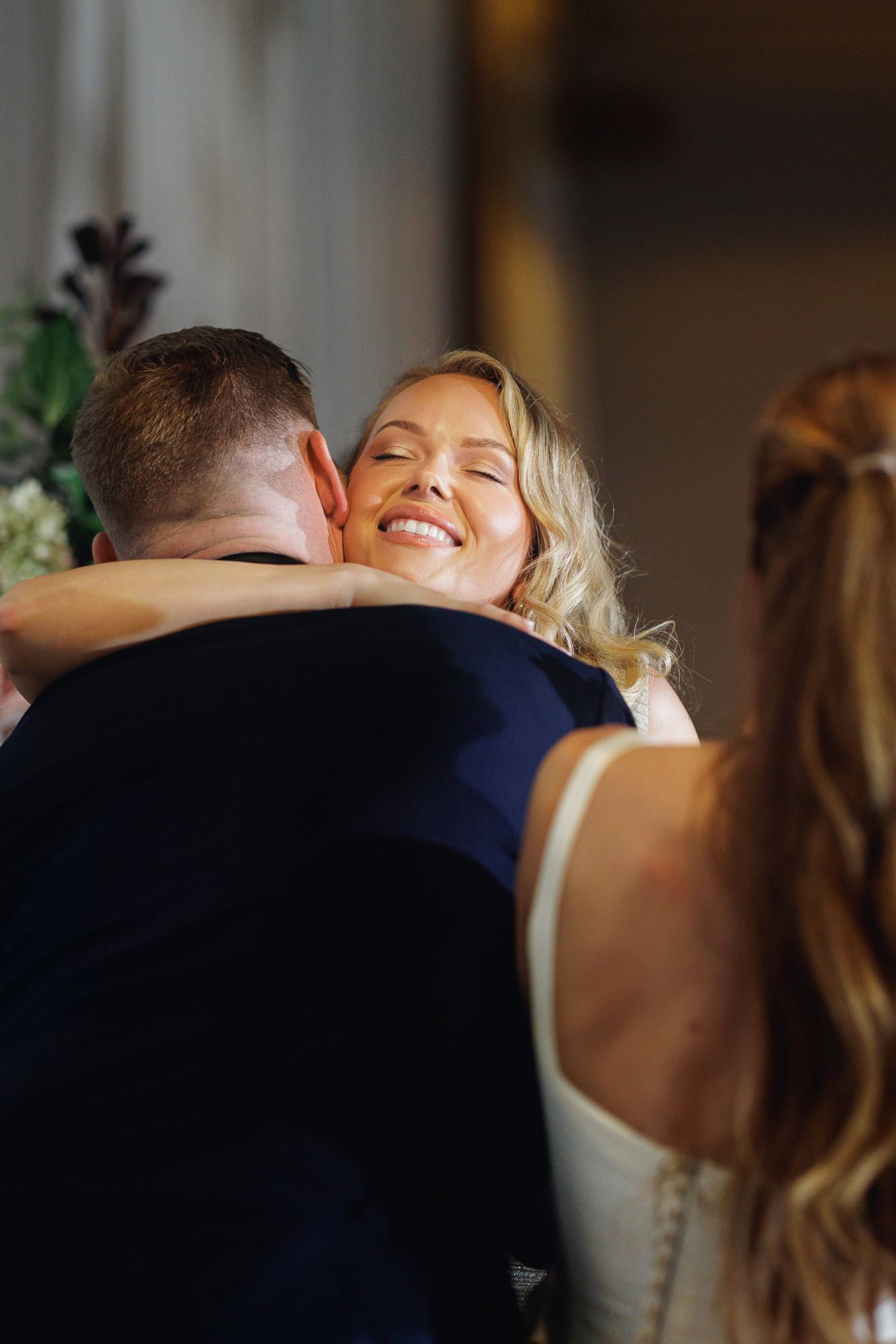 Joyful blonde bride embracing a guest in a navy suit during a warm wedding celebration.