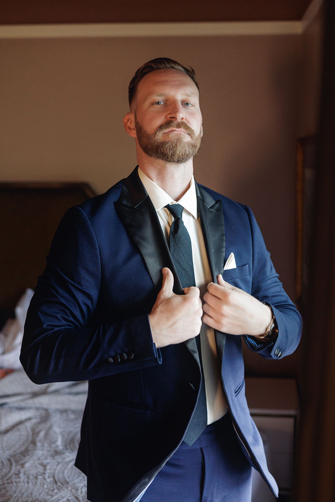 Bearded groom in navy blue tuxedo adjusting lapels, wearing striped tie and white pocket square in hotel room.