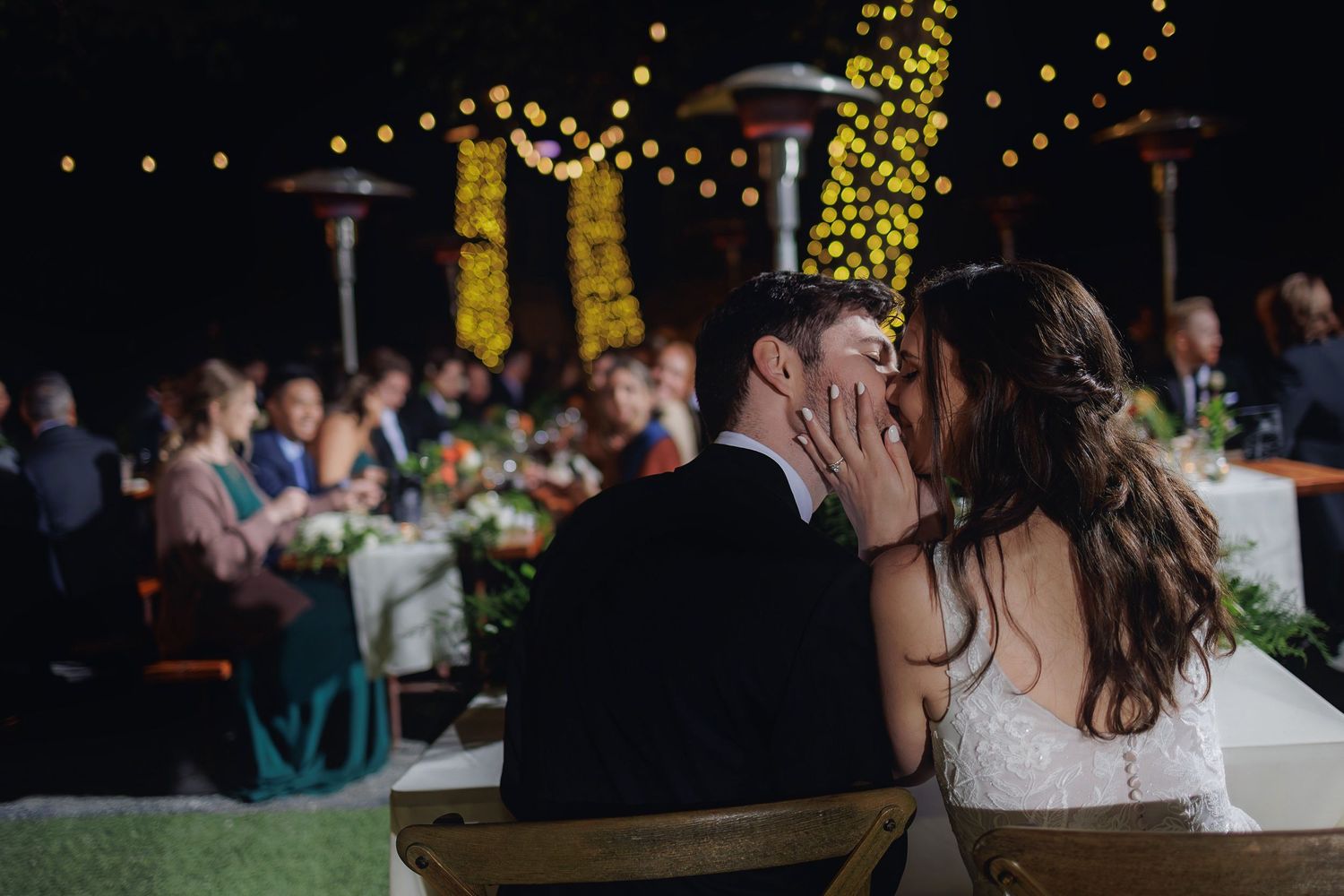 A romantic wedding reception scene outdoors at night with string lights and guests gathered at decorated tables.