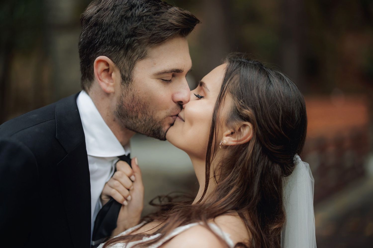 A romantic couple shares a tender kiss outdoors in a blurred natural setting.
