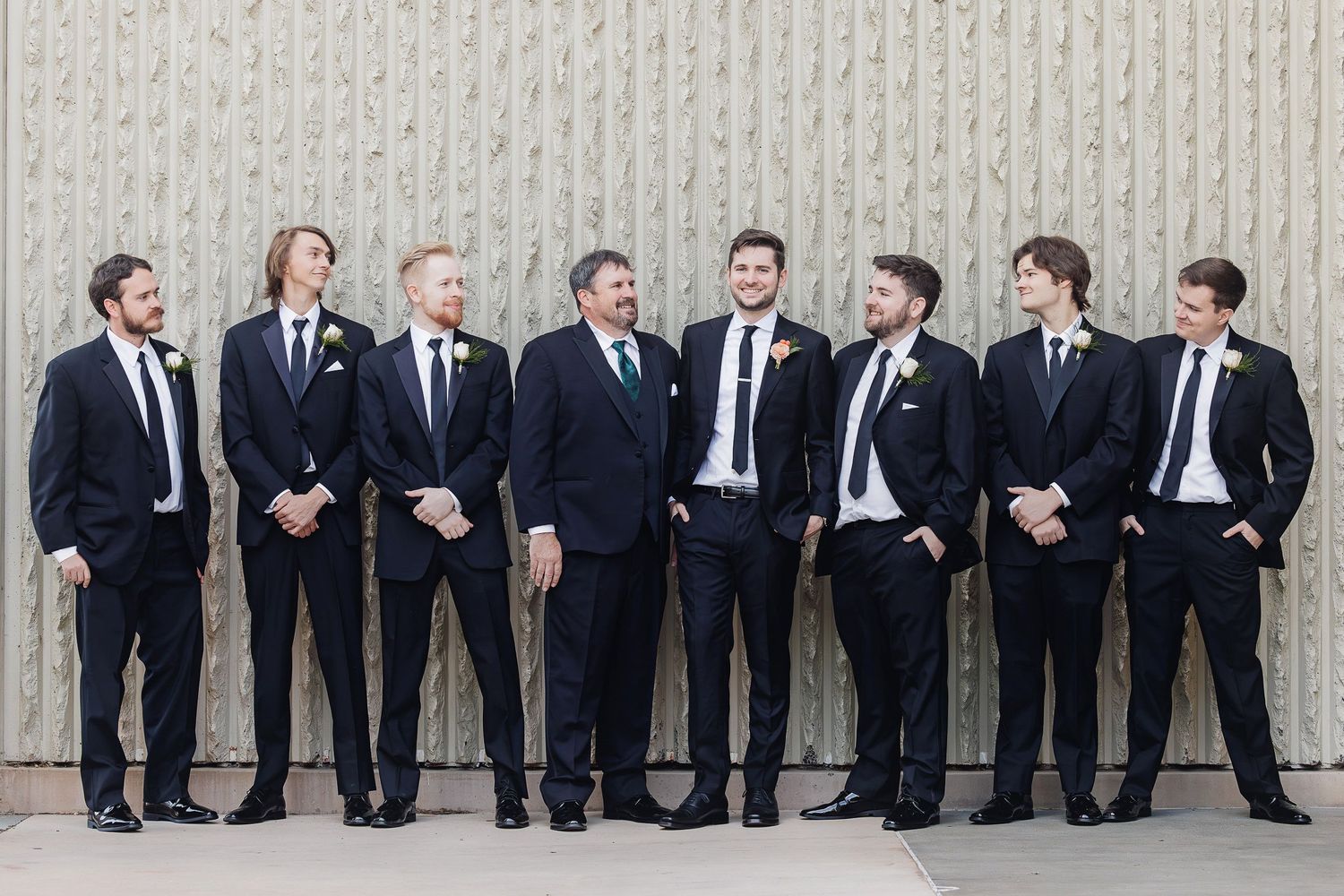 A group of groomsmen in dark suits lean against a white wall sharing laughs and casual conversation.