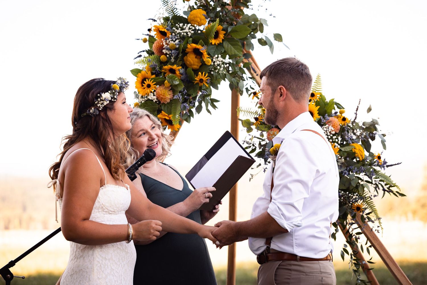 A couple exchanges vows at sunset beneath a floral arch adorned with orange and yellow blooms during an outdoor ceremony.