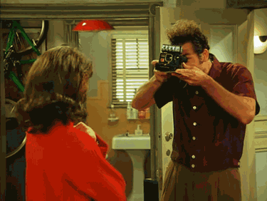 A photographer in a brown shirt takes a picture of someone wearing red in an interior setting.