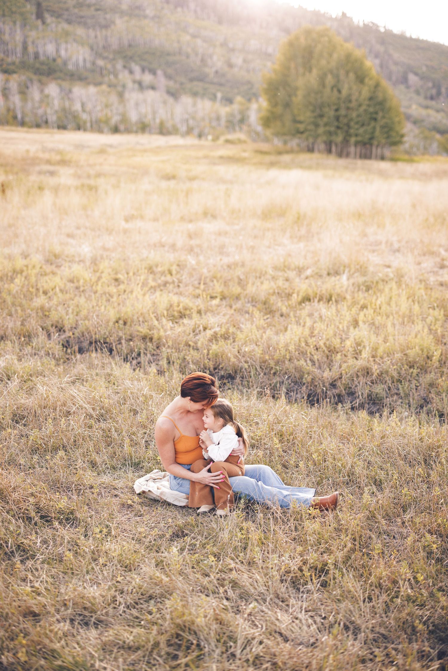 A romantic couple sits together in a sunlit meadow with mountains in the background.